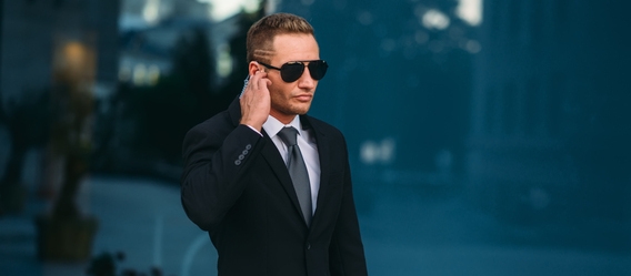 Male bodyguard uses security earpiece outdoors, professional communication tools. Guarding is a risky profession. Guard in suit and sunglasses